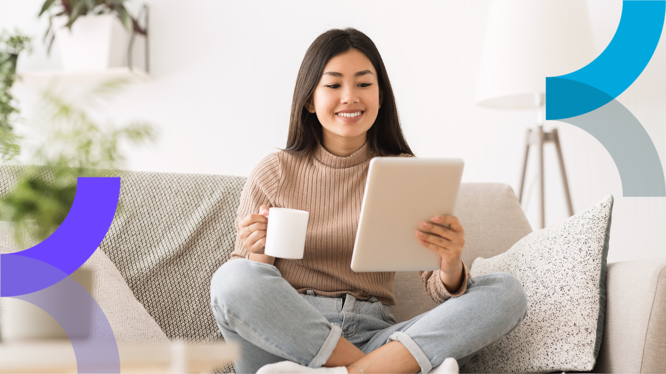 A woman sits on a couch with a mug in her hand and a tablet in the other hand reading consistent marketing campaigns